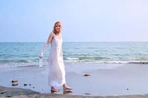 フリー写真 ペットボトルの水を持ってビーチに立つロングワンピ姿の外国人女性